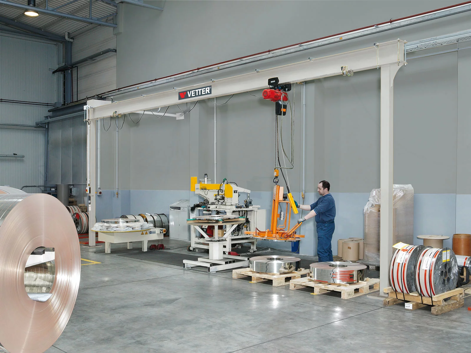 Feststehender ESP-E Einschienen-Portalkran in der Metallverarbeitung. Ein Mitarbeiter hebt Metallringe entlang einer Fertigungslinie mit einem Hebezeug.