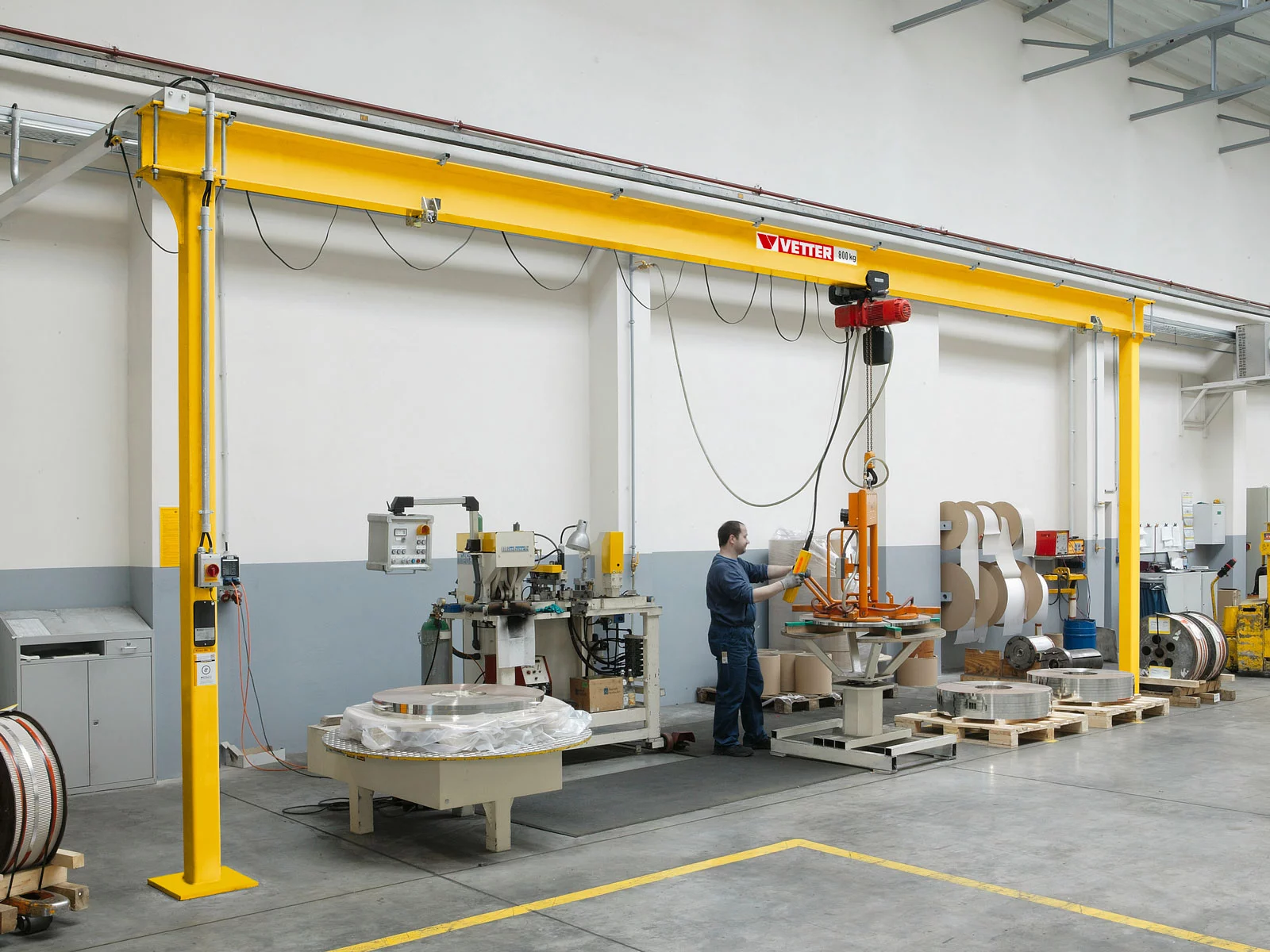 Feststehender ESP-E Einschienen-Portalkran in der Metallverarbeitung. Ein Mitarbeiter hebt Metallringe entlang einer Fertigungslinie mit einem Hebezeug.