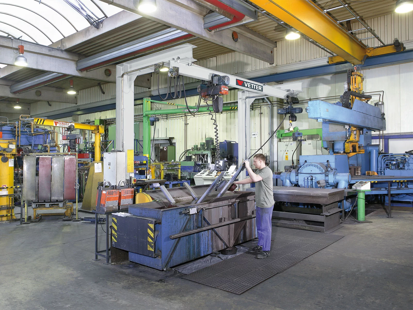 ESP-M Einschienen-Portalkran in lichtgrau in einem metallverarbeitenden Betrieb. In einer Nahaufnahme taucht ein Mitarbeiter Gabelzinken mithilfe des Krans in ein Becken.