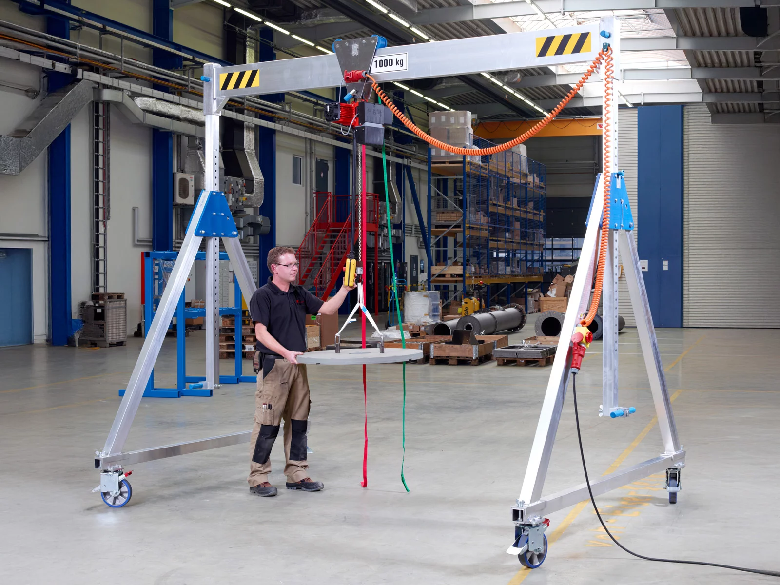 Fahrbarer Aluminium-Portalkran VP-AL in einer Fertigung. Mitarbeiter hebt mit dem Kran einen Betonsockel und bedient das Hebezeug über eine Kommandotafel.