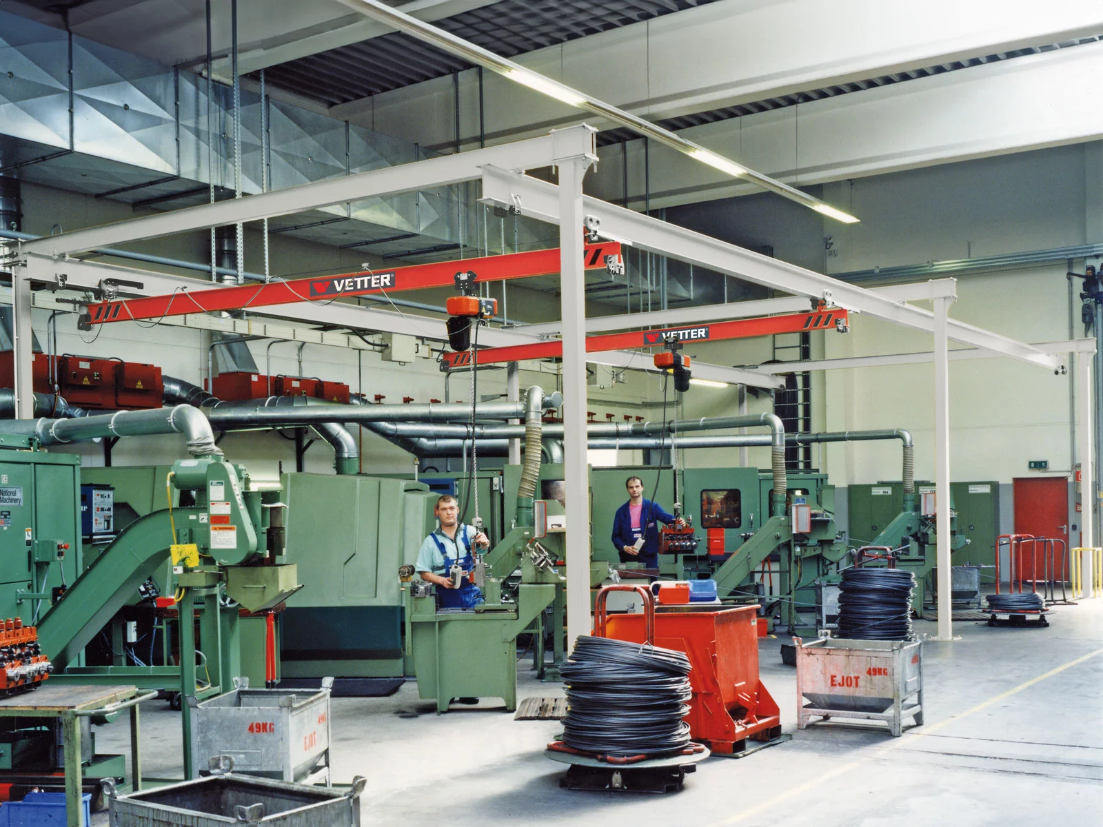ZSP-M Portalkran mit hellgrauen Stützen und roter Brücke in einer Fertigungshalle der Verbindungstechnik. Zwei Mitarbeiter bedienen jeweils eine der Kranbrücken im Portalsystem.