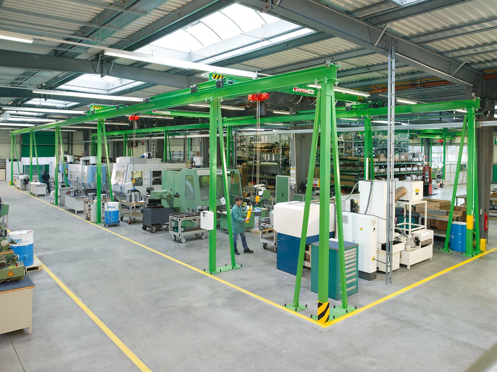 Grüner ZSP-Olympia Portalkran in einer modernen Fertigungshalle, der über Arbeitsplätze verläuft. Ein Mitarbeiter bedient eine Kommandotafel zur Maschinenbeschickung.