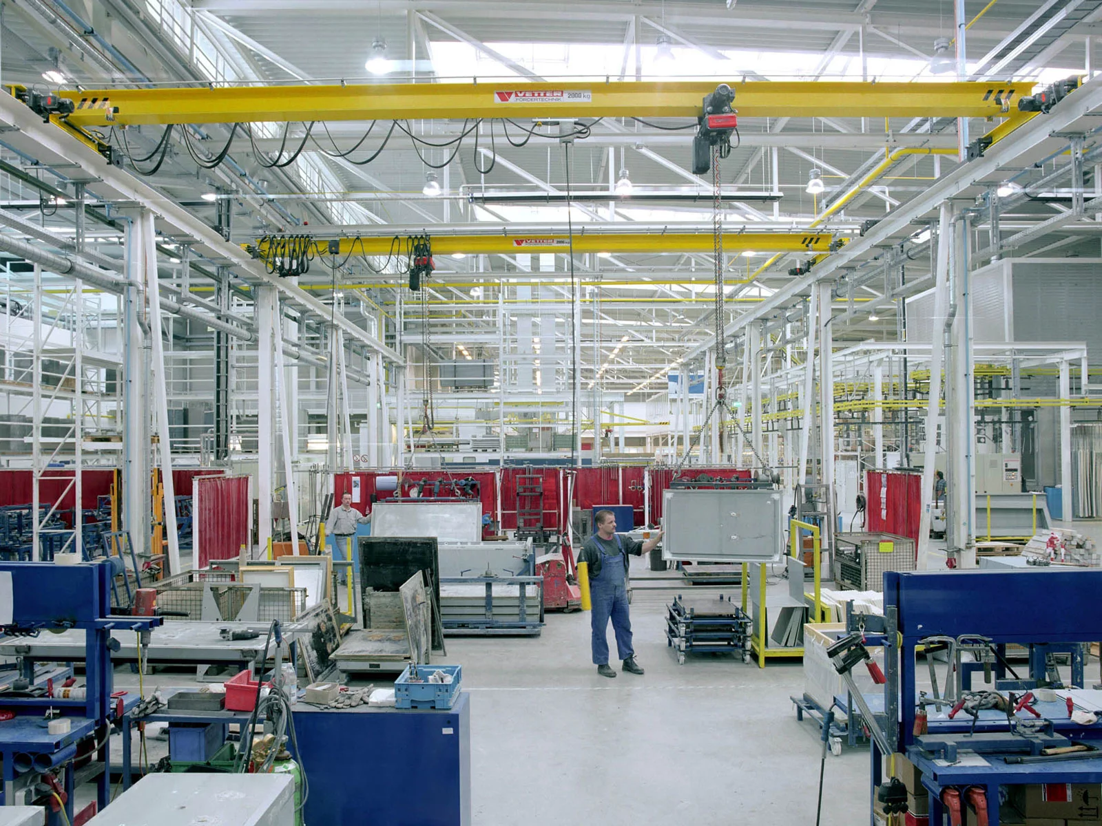 Weißer ZSP-Olympia Portalkran mit gelber Kranbrücke in einer großen Maschinenbauhalle. Ein Mitarbeiter bedient eine Kommandotafel und verfährt das Hebezeug.