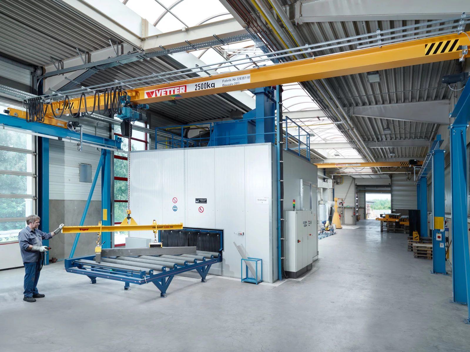 ZSP-Olympia Portalkran mit blauen Stützen und gelber Brücke in einer modernen Industriehalle. Ein Mitarbeiter hebt eine Gabelzinke auf ein Förderband.