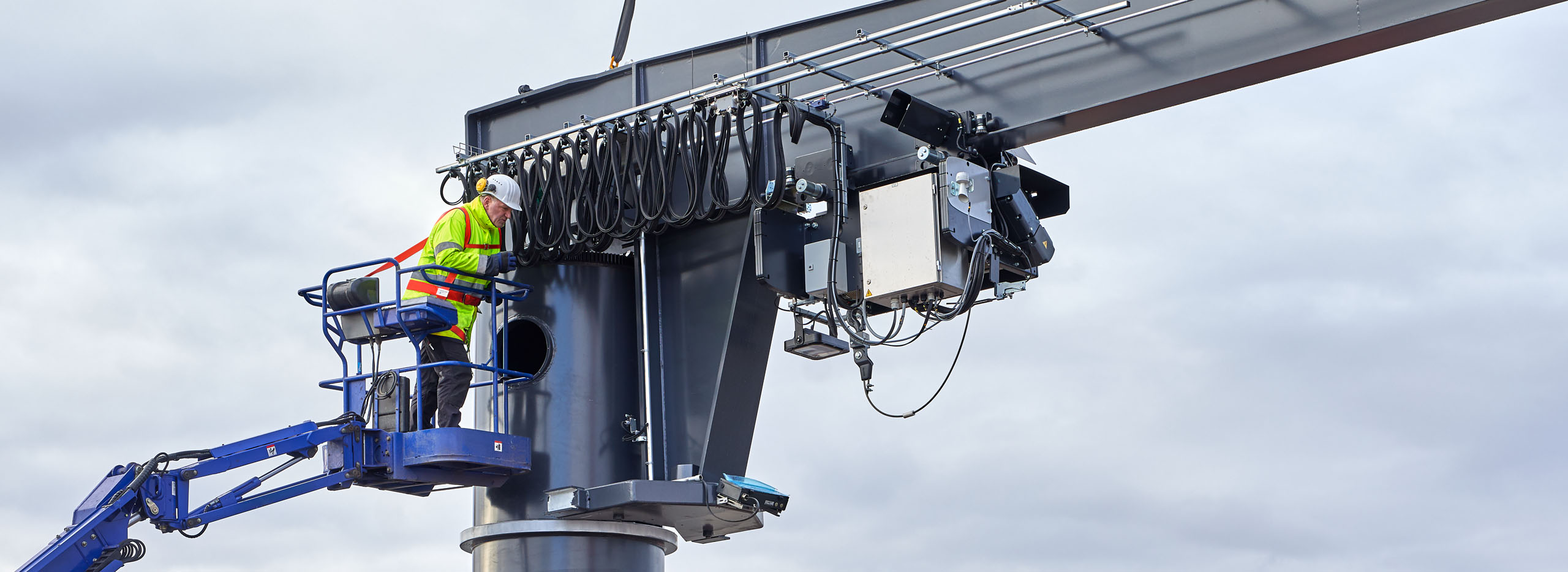 Mitarbeiter in Schutzkleidung montiert Elektronik an grauem Schwenkkran von Hebeb&uuml;hne aus, Kabelbahnhof sichtbar.