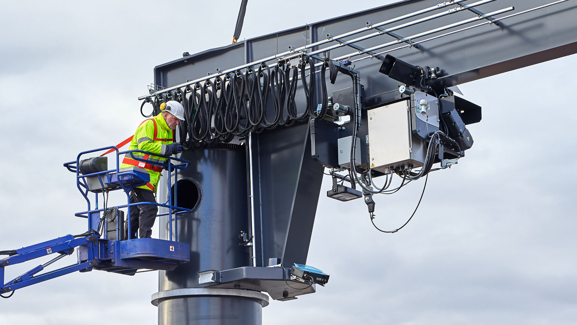 Mitarbeiter in Schutzkleidung montiert Elektronik an grauem Schwenkkran von Hebeb&uuml;hne aus, Kabelbahnhof sichtbar.